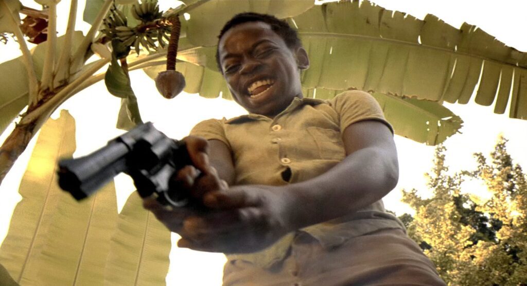 A young boy holding a gun in front of a banana tree, captured in a Dutch Angle Shot.