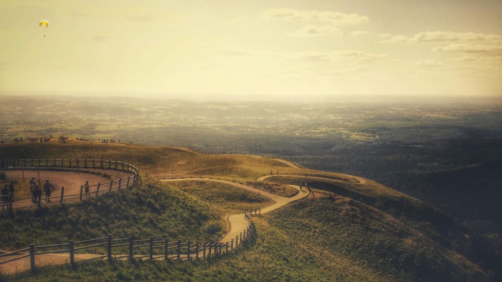 Wide Shot: A person walking down a hill with a parachute in the sky is observed from afar.