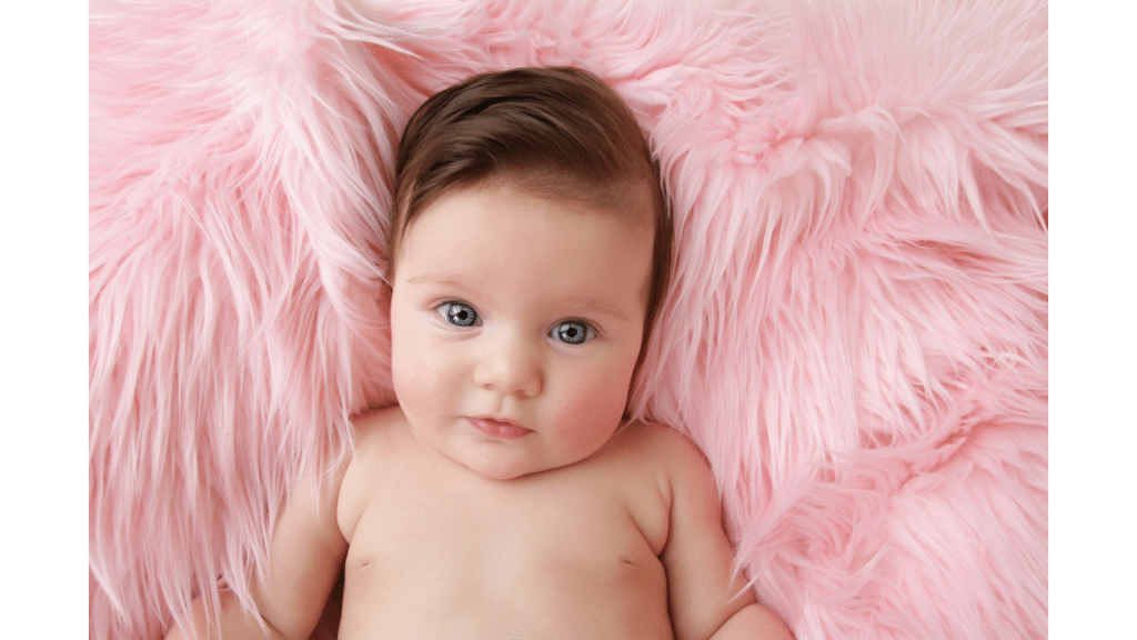 In a display of enchanting newborn photography, a baby with dark hair and blue eyes lies on a pink fluffy surface, gazing directly at the camera.