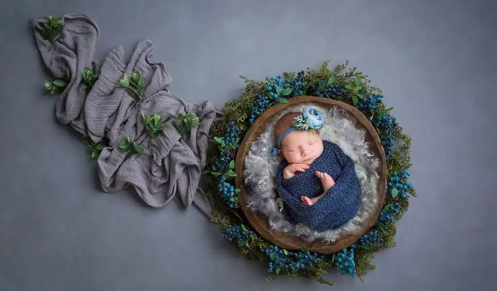 Newborn photography captures a peaceful baby sleeping in a brown basket surrounded by blue flowers and greenery, wrapped snugly in a blue blanket on a gray backdrop adorned with textured purple cloth.