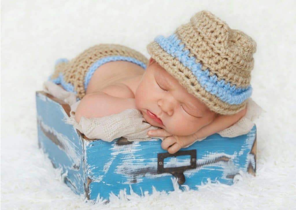 A baby peacefully rests in a small blue wooden box, wearing a knitted hat and shorts, embodying the serene charm of newborn photography against a soft white backdrop.