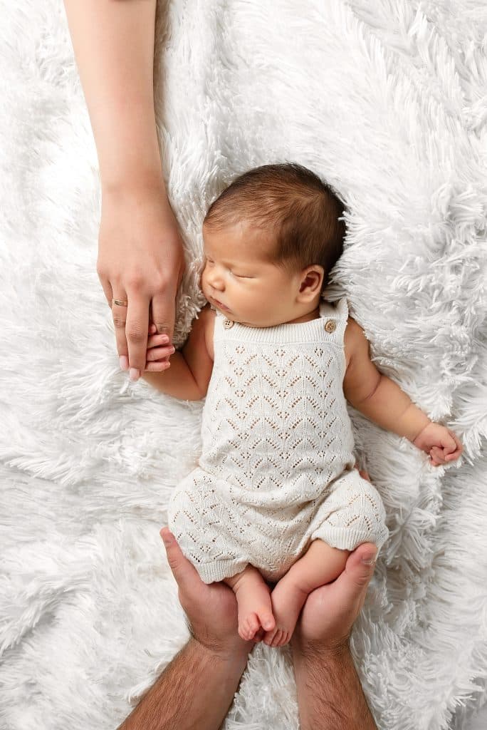 In this tender moment of newborn photography, a baby in a white knit outfit rests on a fluffy white blanket, gently cradled by two adult hands—one softly embracing its legs and the other holding its hand.