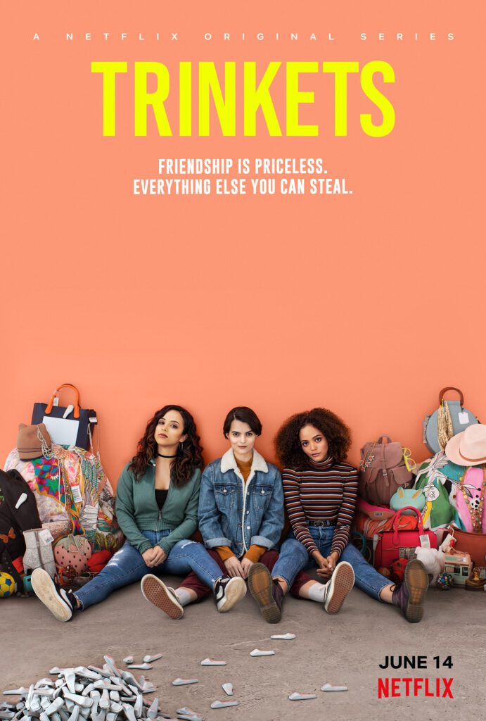 Three young women sitting against a wall with colorful bags and stolen goods around them; text above reads “A Netflix Original Series Trinkets” and “Friendship is priceless. Everything else you can steal.”.