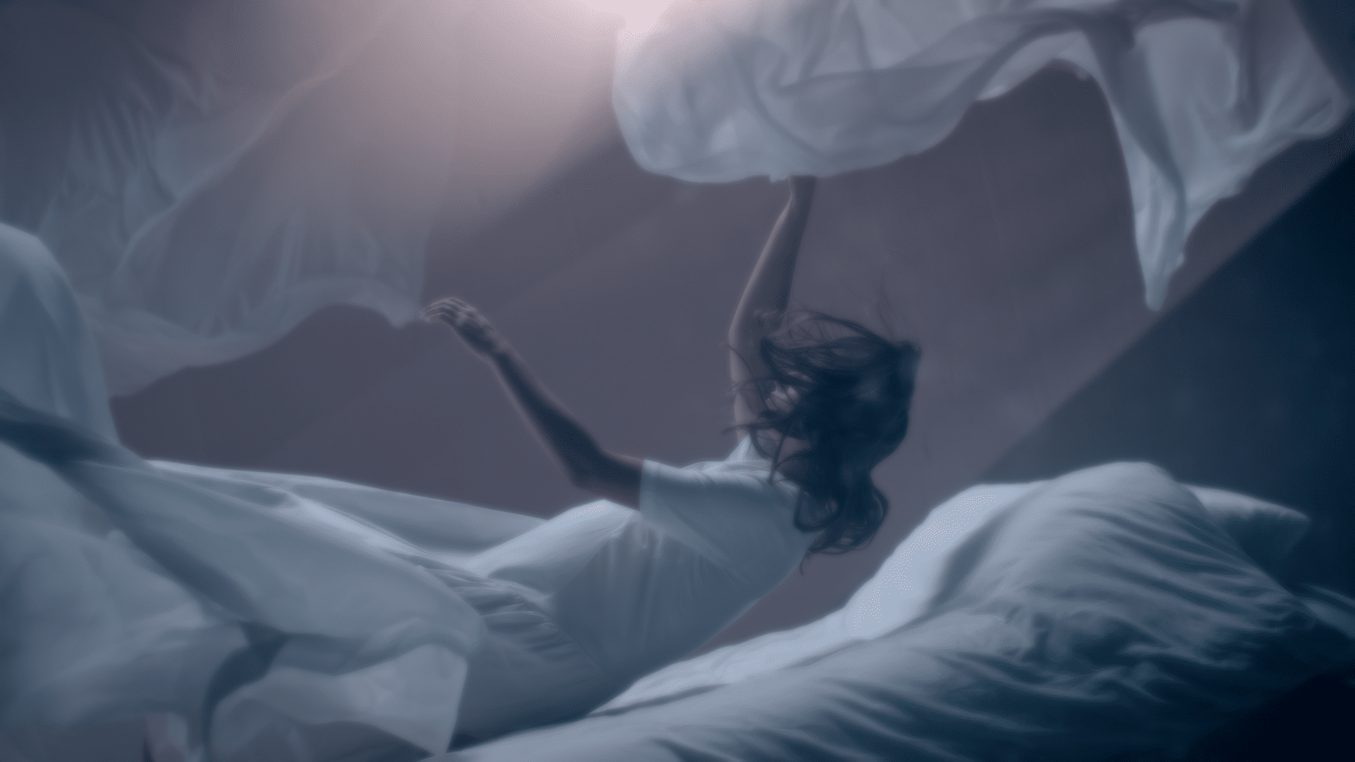 A person in a white dress appears to float above a bed, surrounded by flowing sheets, in a dimly lit room.