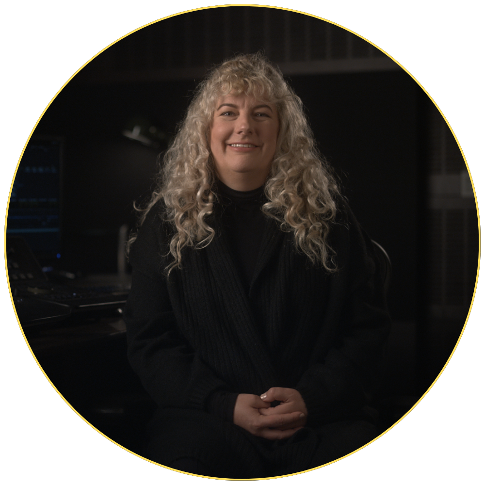 A person with curly hair sits, smiling, in a dimly lit room with a computer in the background.