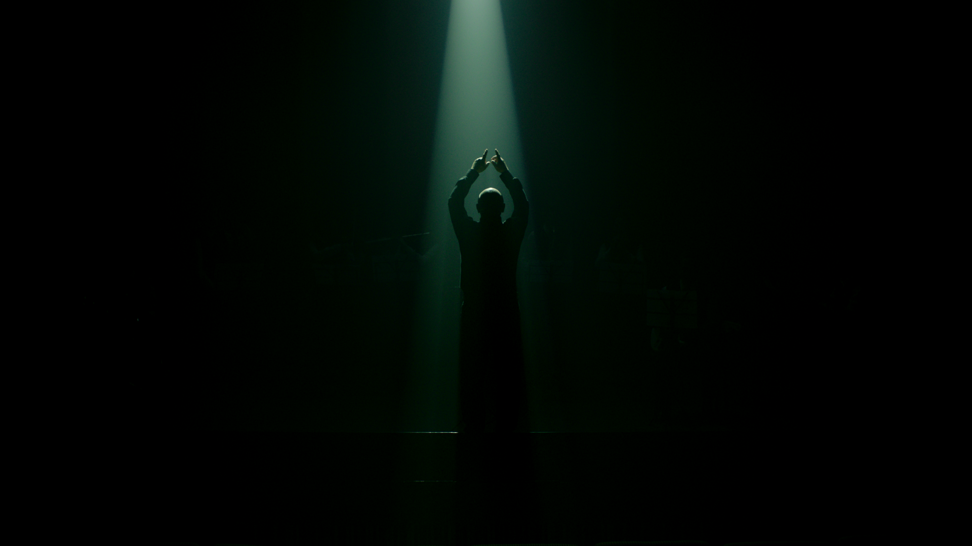 A person stands on stage, silhouetted by a spotlight, with their arms raised.