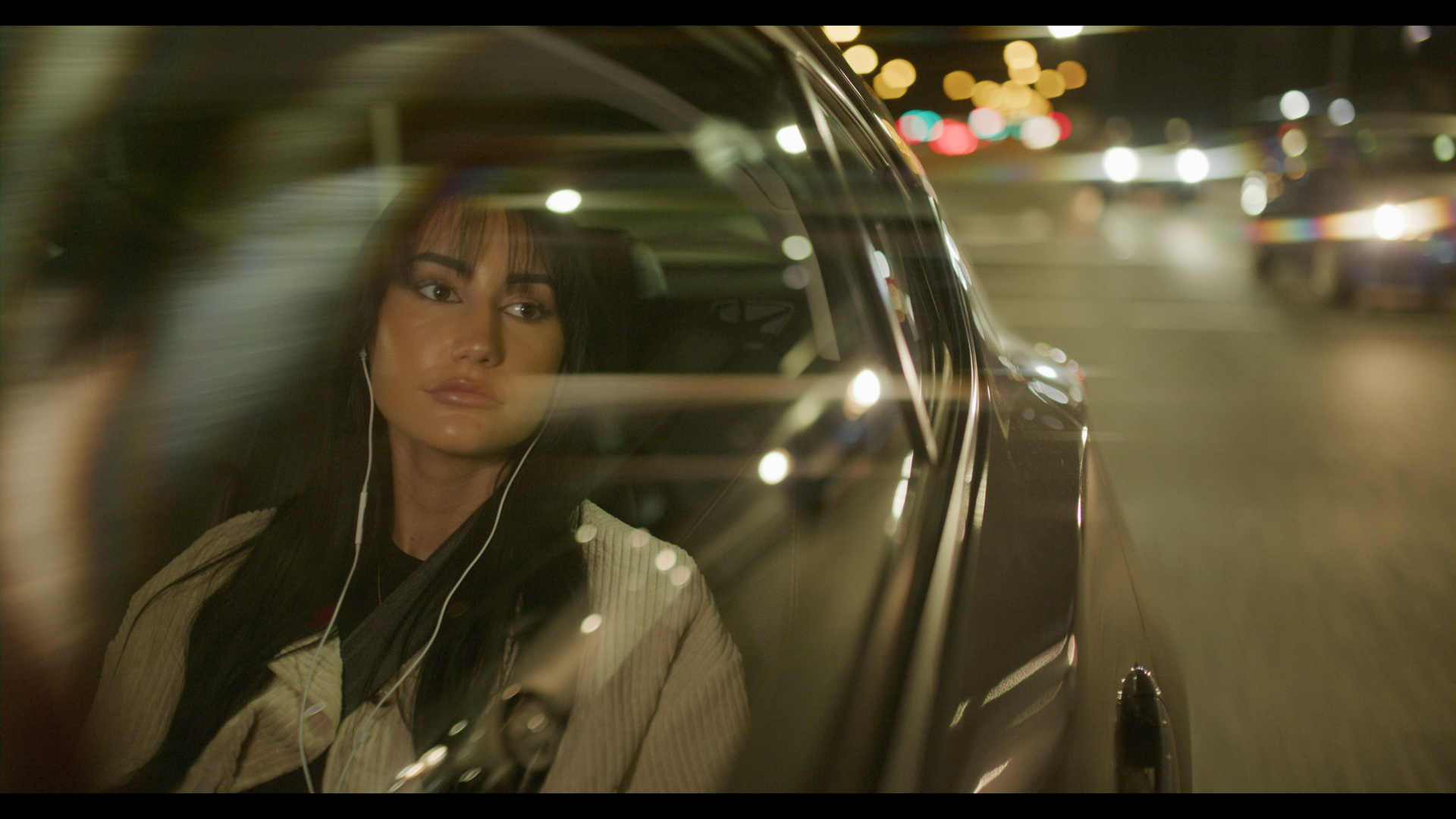 A young woman with earbuds looks out of a car window at night. City lights and streetlights are visible outside.