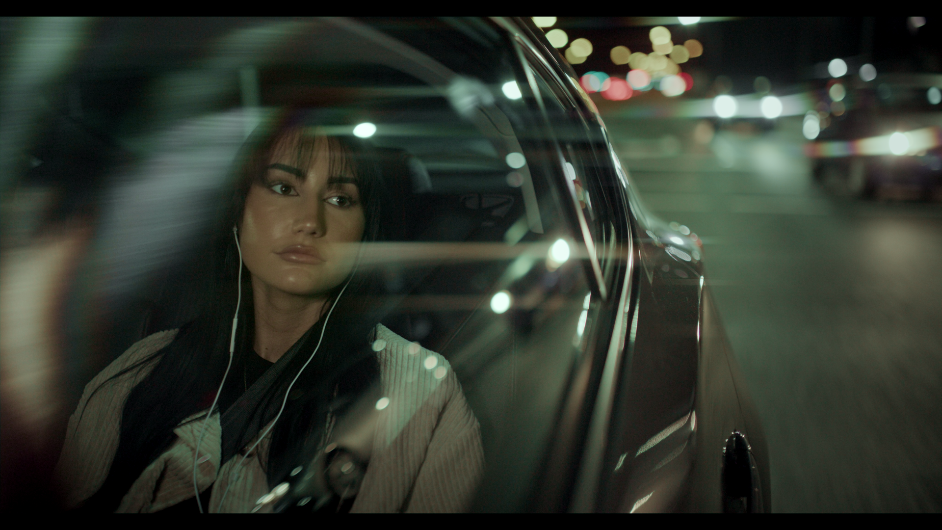 A woman with earphones looks out the window of a moving car at night, with blurred city lights in the background.