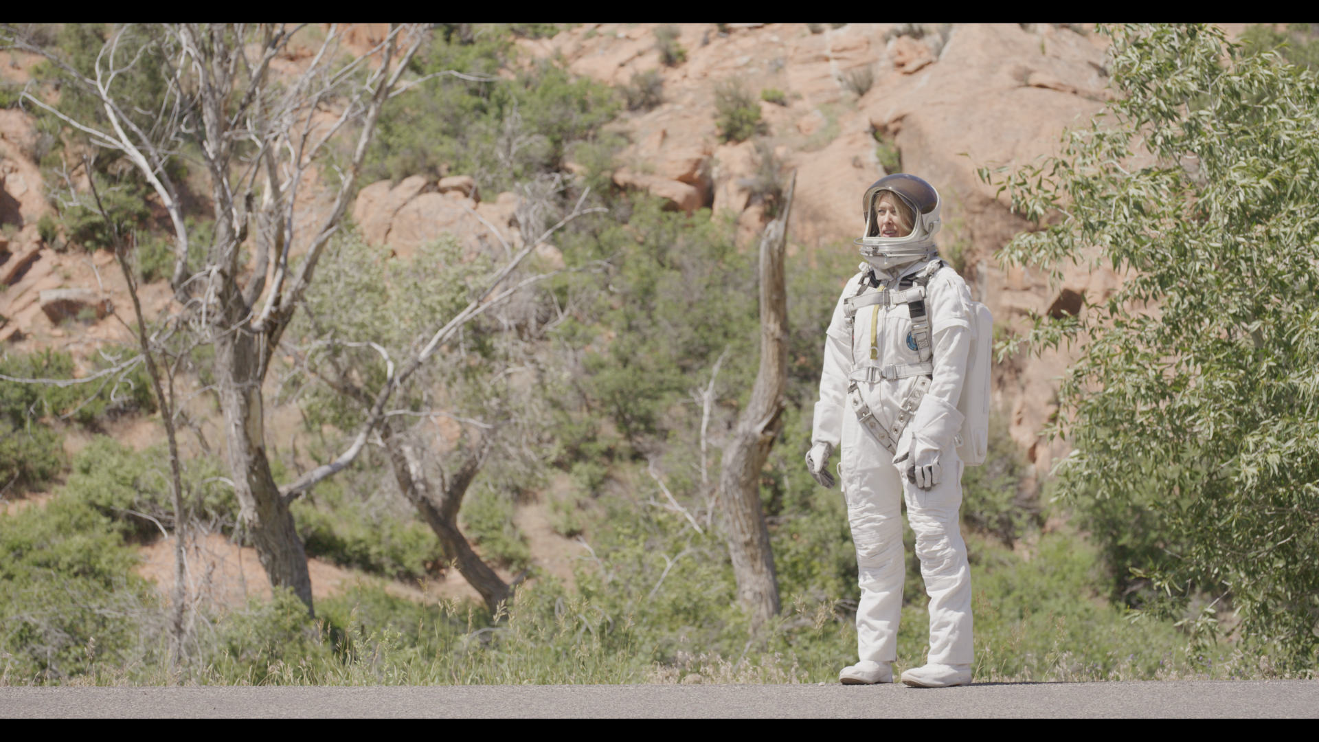 A person in a white spacesuit stands on a road, surrounded by trees and rocky hills in the background.