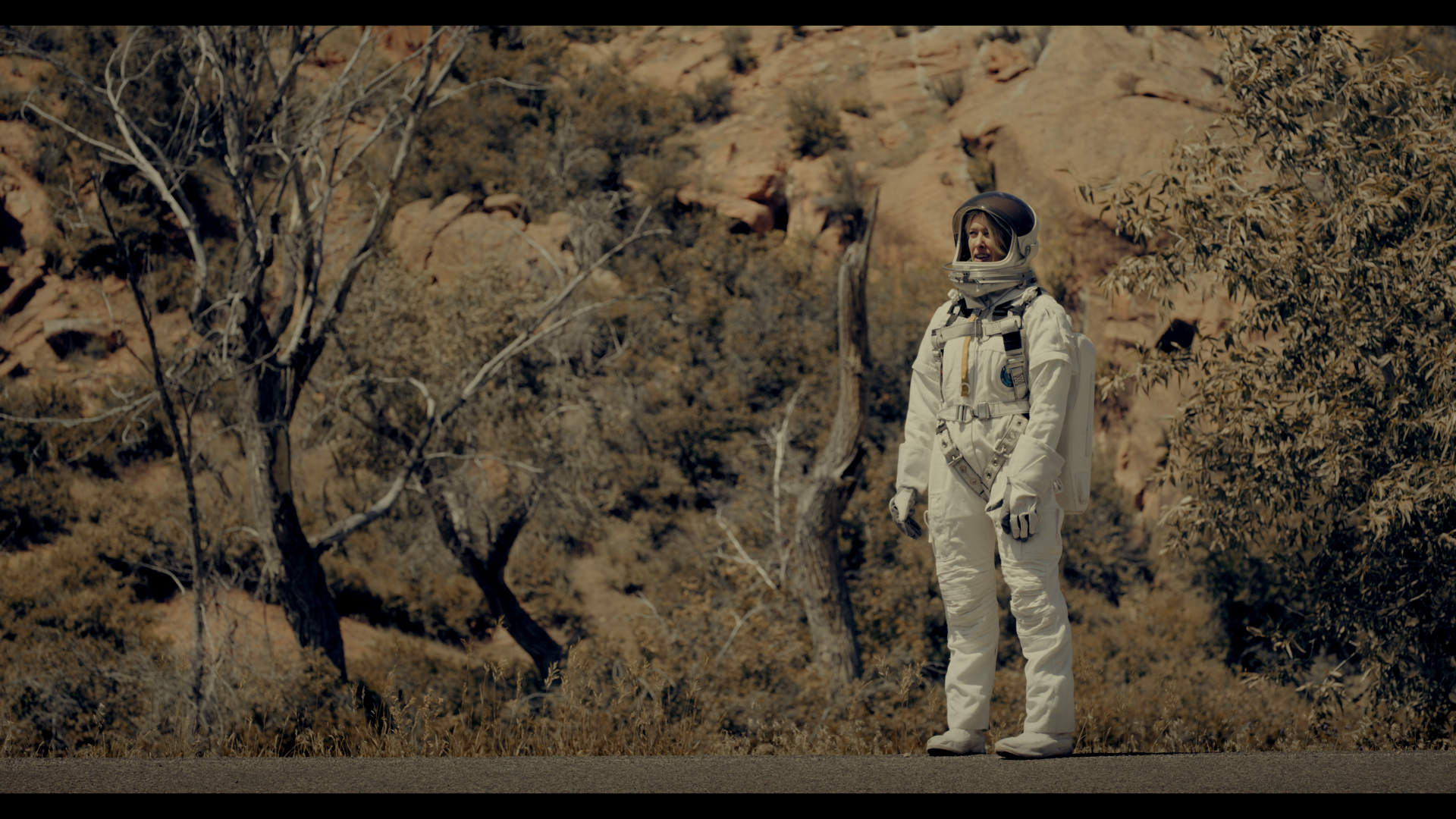 A person in a white spacesuit stands on a road, with a rocky, barren landscape and sparse trees in the background.