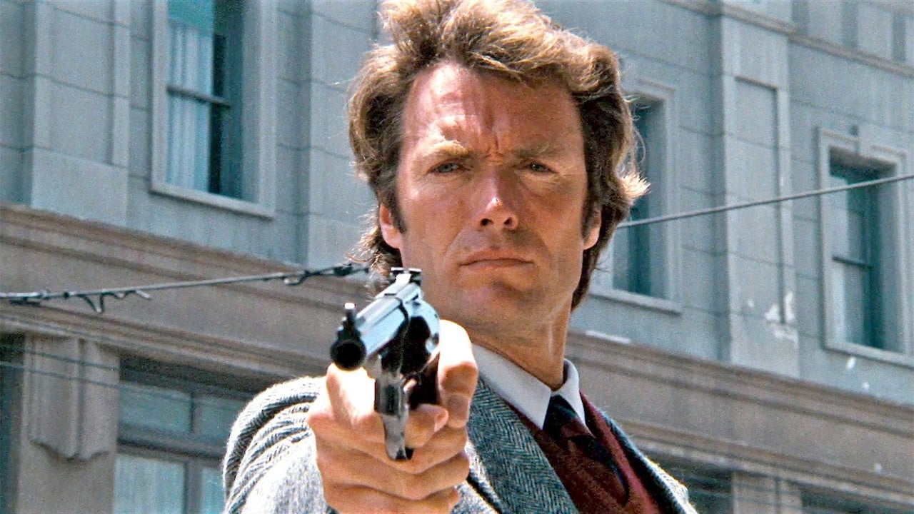 A man in a suit dramatically points a handgun at the viewer, evoking classic scenes of tension typical of guns in movies, with a towering city building set against the backdrop.