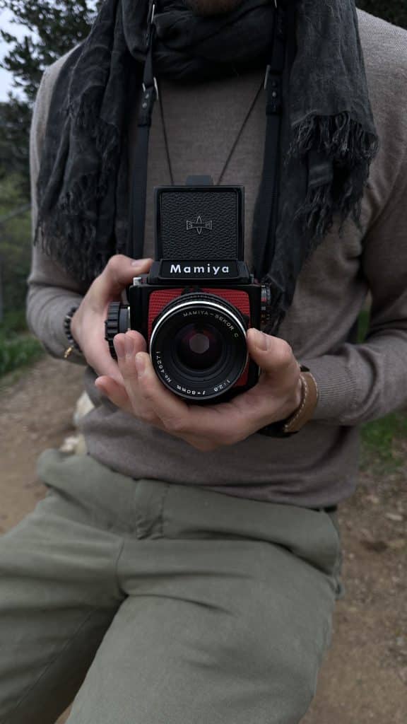 A person stands on a dirt path, holding a Mamiya 645 camera with a red grip, wrapped in a gray scarf and cozy brown sweater.