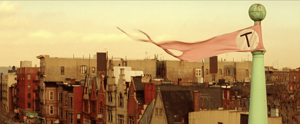 A tattered pink flag with a letter "T" waves atop a tall pole, its hue reminiscent of a Wes Anderson color palette. Urban buildings rise in the background under a cloudy sky, their tones harmonizing with the scene.