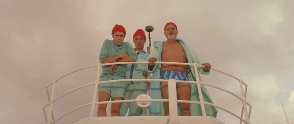 Three individuals in matching red caps and light blue attire stand on a ship's deck, bathed in a Wes Anderson color palette. The man on the right is shirtless and wearing a robe under the overcast sky.