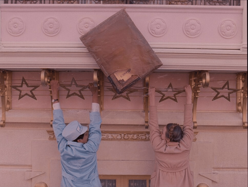 Two people hang from a balcony adorned with golden stars, set against a Wes Anderson color palette. A large brown package labeled "CONFID" tilts precariously above them, adding a quirky yet suspenseful twist to the scene.