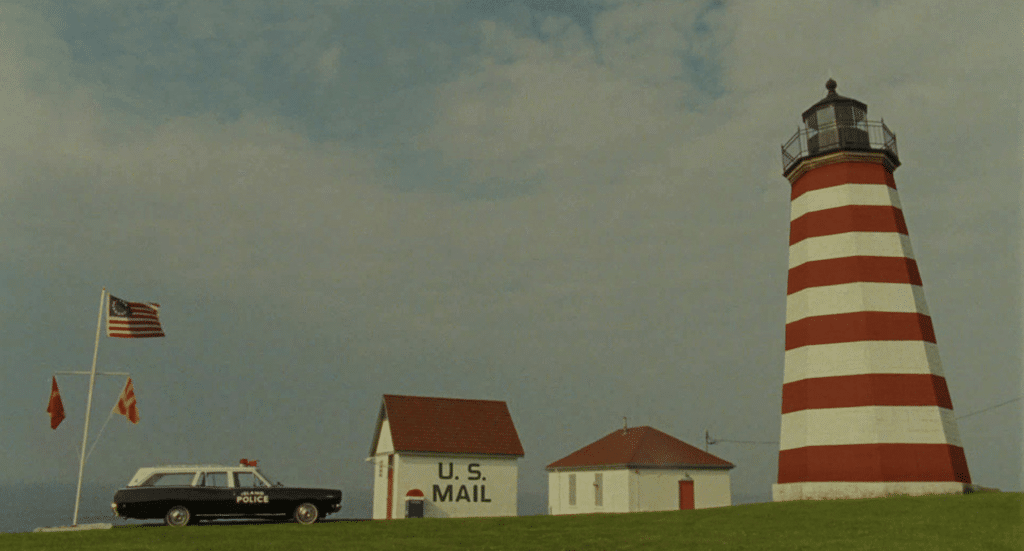 A red and white striped lighthouse towers over a small "U.S. Mail" building with an American flag, while a police car rests on the grass beneath a cloudy sky, all subtly echoing a Wes Anderson color palette.