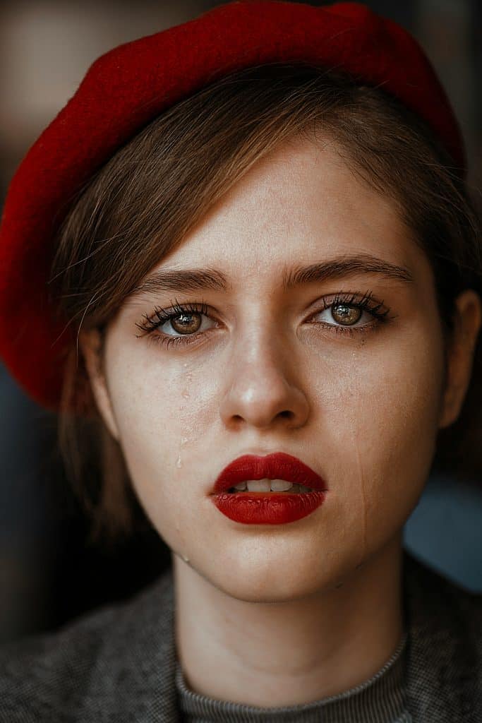 A striking portrait captures a person with short hair, wearing a red beret and bold red lipstick. Tears glisten on their cheeks as they look directly into the camera, conveying raw emotion.