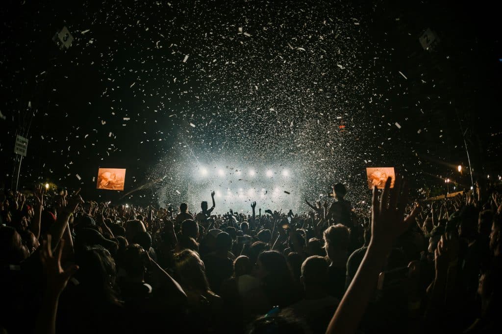 A large crowd at an outdoor concert, with hands raised in a landscape of excitement. Confetti fills the air, and bright stage lights illuminate the background, complemented by vibrant visuals on large screens.