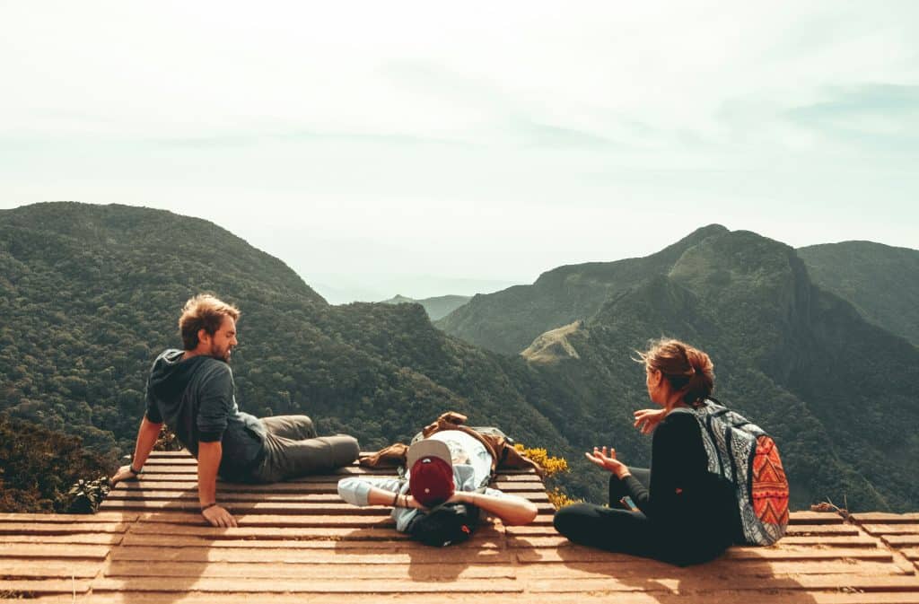 Three people unwind on a wooden platform with a stunning landscape unfolding before them. One person reclines, soaking in the view, while the others sit and chat, their relaxed conversation a portrait of tranquility against the majestic mountains.