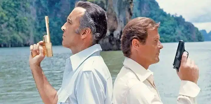 Two men stand back-to-back, each holding a gun like characters from guns in movies. They are outdoors with a body of water and cliffs in the background, both wearing light-colored shirts.