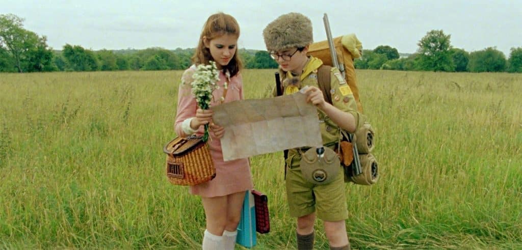 In a grassy field, a girl holds flowers while a boy in a scout uniform, sporting a backpack and coonskin cap, examines a map. The scene unfolds with the distinct charm and whimsical hues of a Wes Anderson color palette.