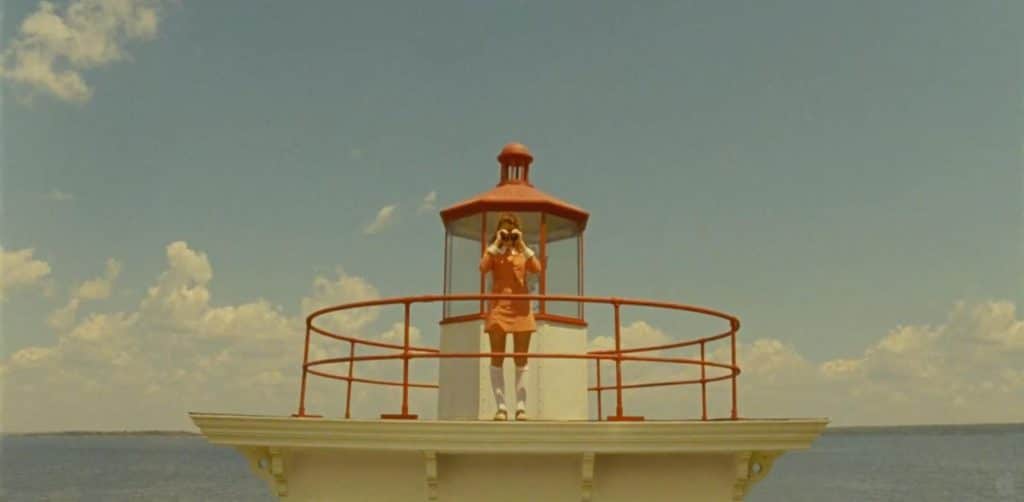 Dressed in orange, the person stands on a lighthouse balcony, gazing through binoculars on a clear day, enveloped by a Wes Anderson color palette that bathes the scene in whimsical pastel hues.