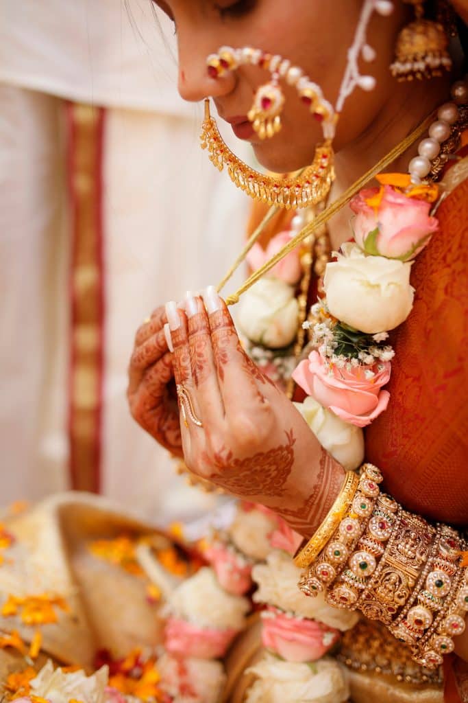 In a portrait of traditional elegance, a person in vibrant attire holds a necklace. Adorned with gold jewelry, floral garlands, and intricate henna on their hands, they exude timeless charm.