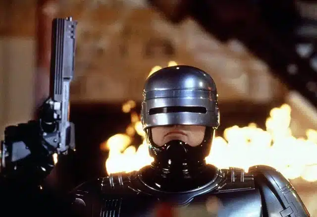 A silver-armored robotic figure holds a pistol, reminiscent of iconic guns in movies, with flames dramatically roaring in the background.