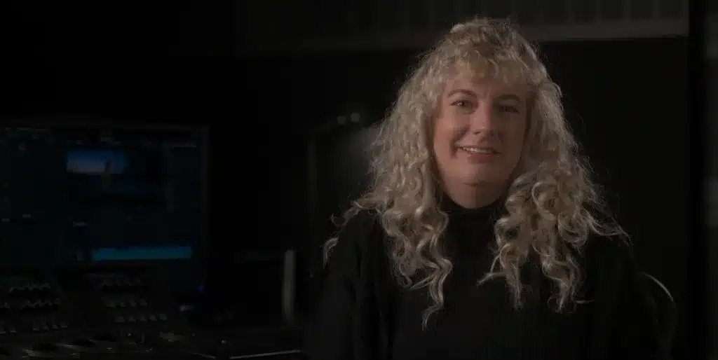 A woman with curly blonde hair and a black sweater sits beside a computer monitor displaying color correction software in a dimly lit room, ready to teach a mini class.